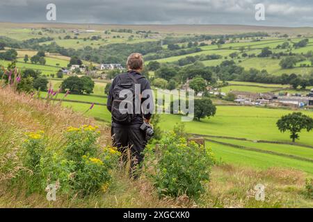 Ein männlicher Walker mit schwarzem Rucksack und Kamera steht am Rand des Baildon Moor in Yorkshire und blickt über Felder im Gill Beck Valley. Stockfoto