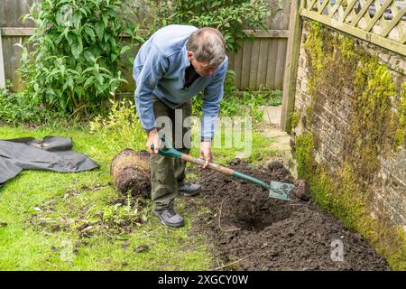 Ein männlicher Gärtner, der ein Loch in ein Blumenbeet gräbt, bereit für das Pflanzen einer großen Pflanze. Der Mann benutzt einen Gartenspaten, um den Boden zu verschieben. Stockfoto