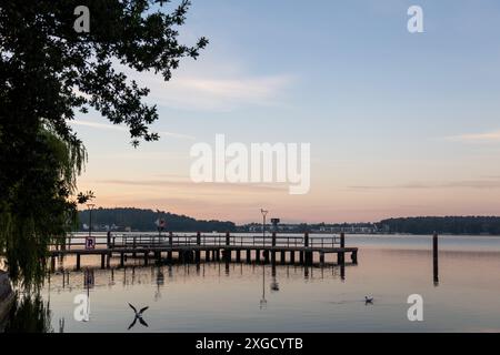 In Waren an der Mueritz, Deutschland, erstreckt sich ein hölzerner Dock mit Geländer in das ruhige Wasser des Sees, während die Sonne hinter einem fernen Wald aufgeht. Stockfoto