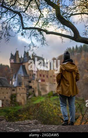 Rückansicht einer jungen Frau mit gelber Jacke, die Eltzer mittelalterliche historische Burg in einer herbstlichen Landschaft mit Bäumen bei Sonnenaufgang, Wierschem, Rhinelan betrachtet Stockfoto