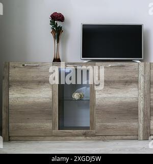 Ein hellbrauner Schrank mit transparentem Glas, darauf ist ein Fernseher, eine Vase mit einer roten Blume, Vorderansicht. Minimalismus-Konzept. Stockfoto