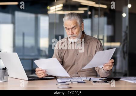 Älterer männlicher Profi, der Papierkram analysiert, während er an einem Laptop in einer modernen Büroumgebung arbeitet. Der Mann konzentriert sich auf die Details der Dokumente und spiegelt Konzentration und Sorgfalt wider. Stockfoto
