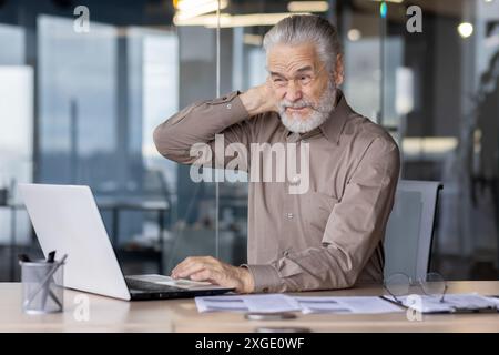 Ein älterer Mann fühlt sich unwohl und Nackenschmerzen, während er einen Laptop an einem Schreibtisch in einer modernen Büroumgebung benutzt. Das Bild vermittelt die Konzepte der Ergonomie am Arbeitsplatz und der alternden Belegschaft Stockfoto