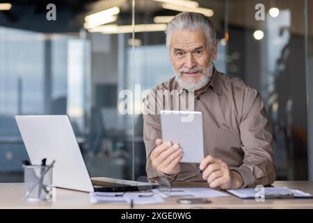 Ein älterer Geschäftsmann verwendet ein Tablet an seinem Schreibtisch, umgeben von einem Laptop und Dokumenten. Der Senior-Mann lächelt selbstbewusst und verkörpert Erfahrung und moderne Technologie in einem professionellen Umfeld. Stockfoto