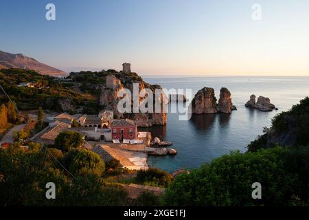 Ein wunderschöner Strand mit einem kleinen roten Gebäude bei Sonnenaufgang. Das Wasser ist ruhig und der Himmel ist wunderschön blau. Tonnara di Scopello, Provinz Trapani, S. Stockfoto