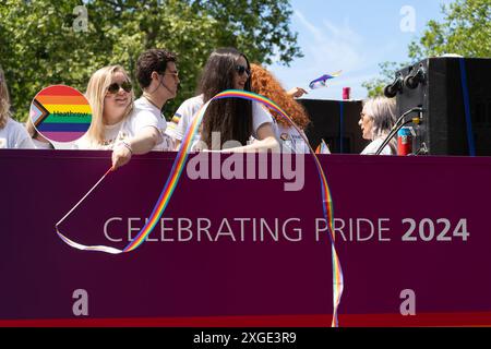 Person, die ein Regenbogenband in einem Heathrow-Bus mit einem Regenbogenlogo und der Botschaft „Celebrating Pride 2024“ auf der London Pride Parade am 29. Juni 2024 winkt Stockfoto