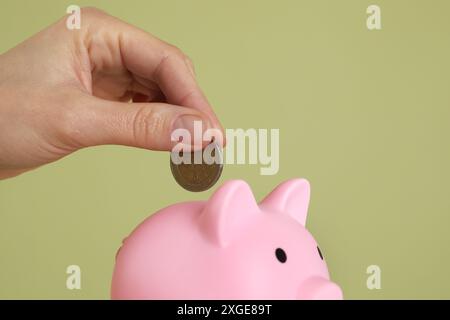 Frau, die Münze in rosa Schweinebank auf olivfarbenem Hintergrund steckt, Nahaufnahme. Leerzeichen für Text Stockfoto