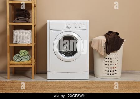Waschmaschine, Korb und Regal in der Nähe beigefarbener Wände im Innenbereich Stockfoto