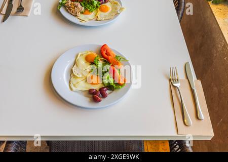 Zwei Teller Frühstück mit sonnigen Eiern, frischem Salat, Tomaten und Oliven auf weißem Tisch, Besteck und Servietten neben Tellern. Stockfoto