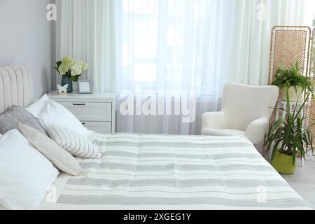 Großes Bett, Kommode, Sessel, Zimmerpflanzen und Fenster mit Vorhängen im Schlafzimmer. Innenarchitektur Stockfoto