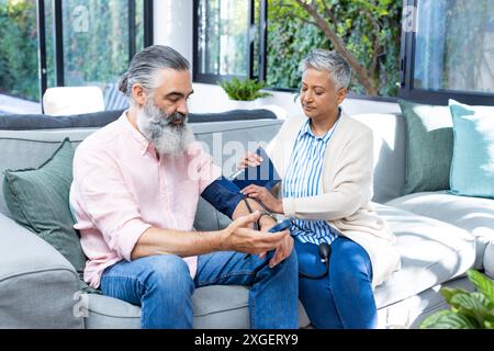 Ältere Frau, die den Blutdruck eines Mannes überprüft, der zu Hause auf der Couch sitzt Stockfoto