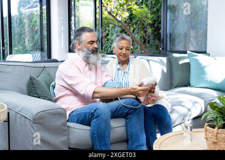 Senior Pärchen, die den Blutdruck mit dem Monitor überprüfen, während sie auf der Couch sitzen Stockfoto