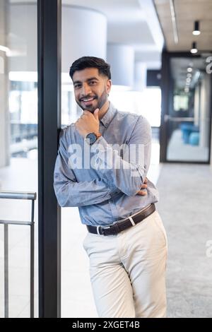 Lehnt sich an eine Glaswand, lächelnder Geschäftsmann im Büro in eleganter Freizeitkleidung Stockfoto