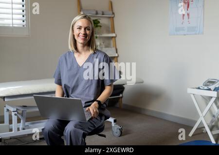Lächelnde Krankenschwester in Schrubben mit Laptop in der Arztpraxis, auf Hocker sitzend, Kopierraum Stockfoto