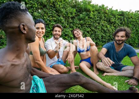 Gruppe von Freunden, die sich auf dem Gras entspannen - einen Sommergarten genießen - geselliges Treffen im Freien, verschiedene Gruppe von Freunden - plaudern und lachen, re Stockfoto
