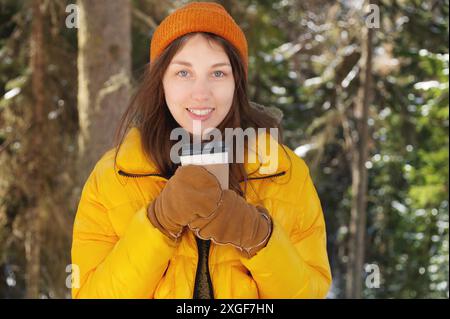 Attraktive kaukasische positive junge Frau in Winterkleidung mit Hut mit Fäustlingen und Daunenjacke steht in einem Nadelwald mit Papier Stockfoto