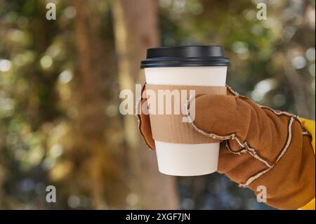 Nahaufnahme einer weiblichen Hand in einem Fäustling, die eine Papiertasse mit Kaffee oder Tee im Freien in einem winterlichen Nadelwald hält Stockfoto