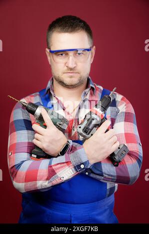 Seriöse kaukasische Werkstatt in Brillen mit Holzbearbeitungswerkzeugen. Zimmermannsporträt auf rotem Studiohintergrund Stockfoto