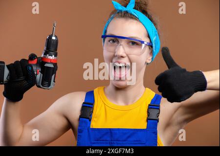 Attraktive junge kaukasische Frau in Arbeitsuniform mit Schutzbrille und einem Schraubenzieher in der Hand, der glücklich lächelt und die Daumen-nach-oben-Geste auf Braun zeigt Stockfoto
