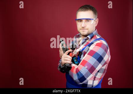 Ernsthafter kaukasischer Werkstattmeister in Brille mit Holzbearbeitungswerkzeugen. Zimmermannporträt auf rotem Atelierhintergrund. Kopierbereich Stockfoto