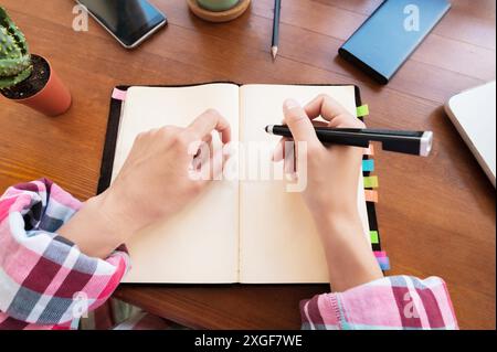 Junge weiße attraktive Frau macht Notizen in ihrem Notizbuch, während sie in der Küche am Tisch sitzt. Notizbuch aus Papier, das weibliche Hände schreibt Stockfoto