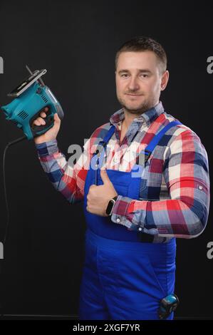 Ein lächelnder Mann in einem karierten Hemd und Overall hält eine Puzzle in der Hand. Auf schwarzem Hintergrund und mit der anderen Hand zeigt eine Daumen-nach-oben-Geste Stockfoto