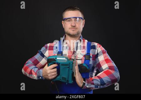 Ein lächelnder Mann in einem karierten Hemd und Overall hält eine Puzzle in der Hand. Auf schwarzem Hintergrund und mit der anderen Hand eine Namaste-Geste Stockfoto