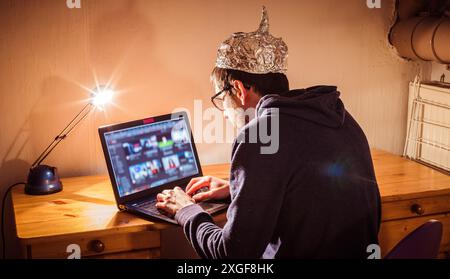 Der junge Mann mit Aluminiumkappe sitzt im dunklen Keller vor einem Laptop. Verschwörungstheorie Konzept Stockfoto