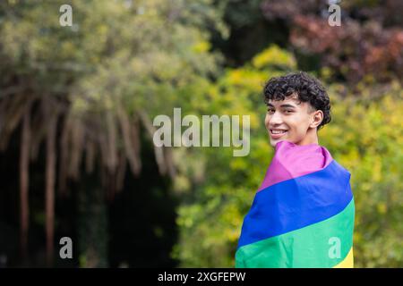 Ein lächelnder junger Mann in Regenbogenfahne, der im Freien den LGBTQ+-Stolz feiert, Kopierraum Stockfoto