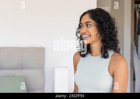 Lächelnde Frau mit lockigem Haar, die im Schlafzimmer steht, wegblickt, Kopierraum Stockfoto