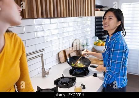 Gemüse im Wok kochen, Frau lächelt und spricht mit einem Freund in der Küche Stockfoto