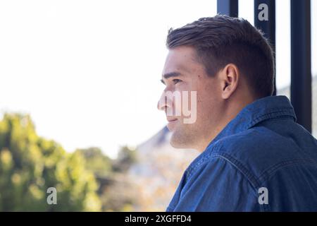 Junger Mann in Denim-Hemd, der nachdenklich aus dem Fenster blickt, den Blick auf die Natur genießt, den Kopierraum Stockfoto