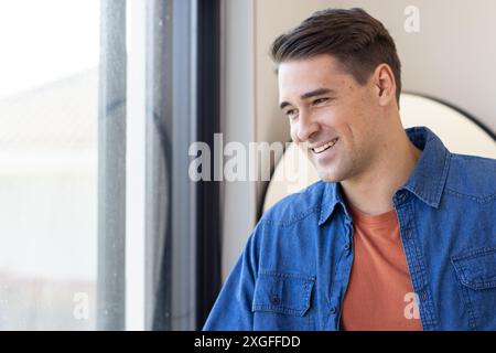 Lächelnder Mann in lässigem Jeansshirt, der am Fenster steht und nach draußen blickt Stockfoto