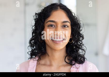 Lächelnde Frau mit lockigen Haaren, die ein rosa Hemd trägt und drinnen in die Kamera schaut Stockfoto
