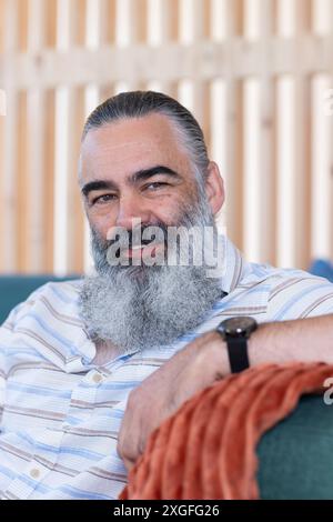 Lächelnder, bärtiger Seniorenmann, der sich auf der Couch entspannt und die Freizeit drinnen genießt Stockfoto