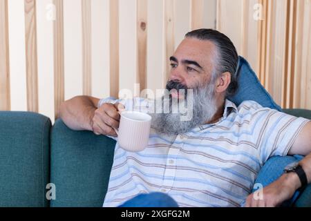 Entspannen Sie auf dem Sofa, ein älterer Mann mit Bart trinkt Kaffee und genießen Sie die Freizeit Stockfoto