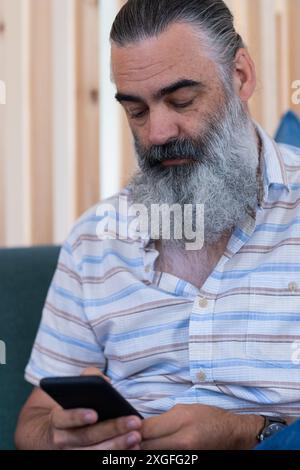 Mit dem Smartphone konzentriert sich der bärtige Seniorenmann, während er drinnen auf der Couch sitzt Stockfoto