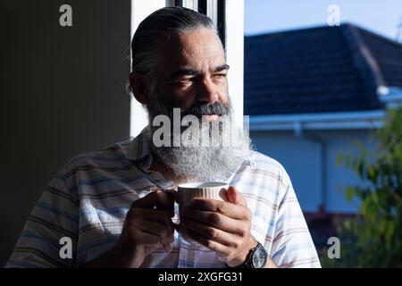 Mit Becher, Seniorenmann mit Bart, der drinnen das Morgensonnenlicht genießt Stockfoto