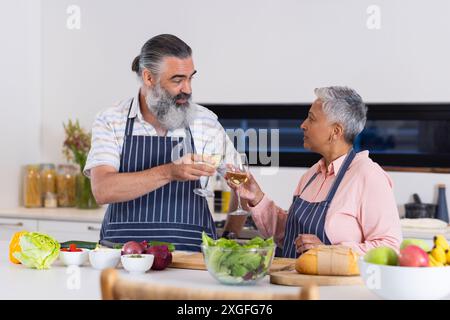 Seniorenpaar, das mit Weingläsern tostet, während Salat in der Küche zubereitet wird Stockfoto