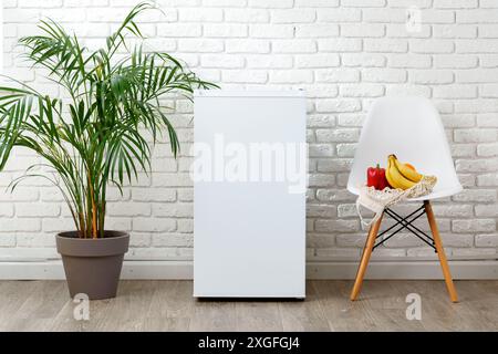Kühlschrank gegen weiße Ziegelwand mit grüner Palmenpflanze und weißem Stuhl mit Obst Stockfoto