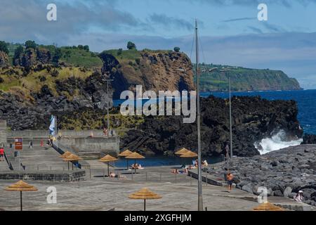 Eine felsige Küste mit Menschen in einem Meerwasserpool, die unter Sonnenschirmen ruhen, während das Meer gegen die Felsen schlägt, Capelas, Sao Miguel Island Stockfoto