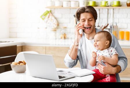 Glücklicher Vater Spricht Am Telefon, Während Er Das Baby In Der Küche Hält Stockfoto