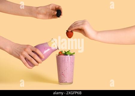 Frau, die frischen Smoothie aus der Flasche in Glas gießt, und weibliche Hände mit Beeren auf gelbem Hintergrund Stockfoto