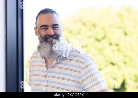 Lächelnder älterer Mann mit grauem Bart, der draußen in einem lässigen gestreiften Hemd steht Stockfoto