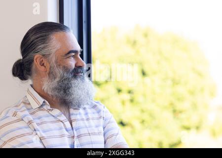 Lächelnder älterer Mann mit grauem Bart, der aus dem Fenster blickt, einen friedlichen Moment genießt, Kopierraum Stockfoto