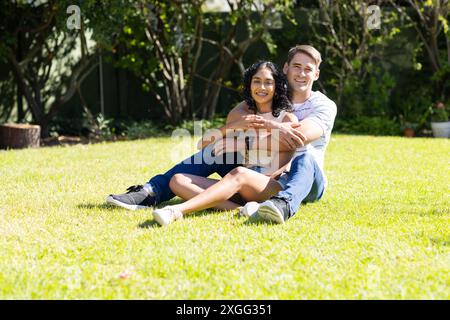 Auf Gras sitzend, junges Paar umarmt und lächelt im sonnigen Garten Stockfoto