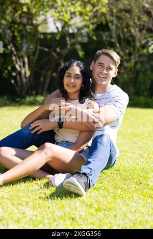 Ein junges Paar, das im Park auf Gras sitzt, umarmt und lächelt in die Kamera Stockfoto