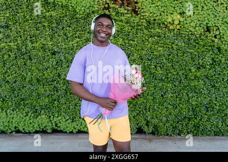 Ein afrikanischer Mann, der ein lila Hemd trägt, hält einen Blumenstrauß. Er trägt Kopfhörer und hört Musik Stockfoto