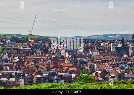 Ein Panoramablick auf eine Stadt mit roten Häusern, einem Kran im Hintergrund und grünen Hügeln in der Ferne unter einem bewölkten Himmel, Whitby, Yorkshire Stockfoto