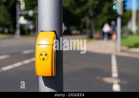 Gelbe Taste mit drei schwarzen Punkten am grauen Ampelmast. Hintergrund zeigt Straße, Bürgersteig mit nicht fokussierten Menschen, die gehen. Stockfoto
